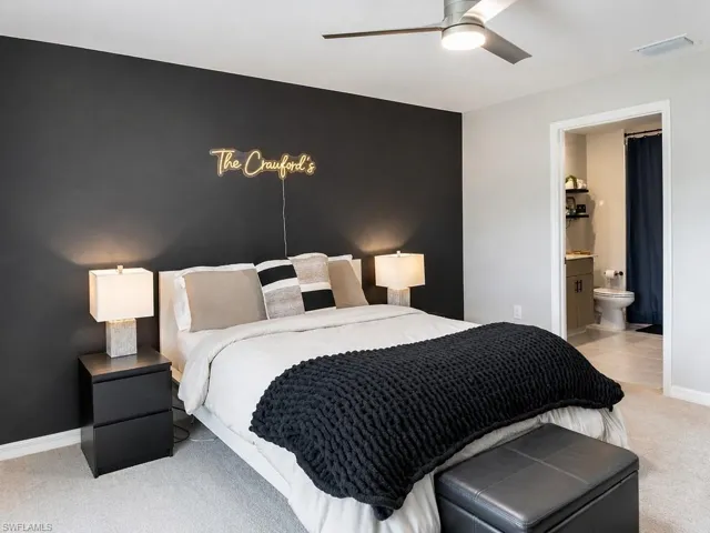 Carpeted sleeping area featuring a dark accent wall with overhead lighting, a ceiling fan, and an open doorway revealing a bathroom with tile flooring and dual sink vanity