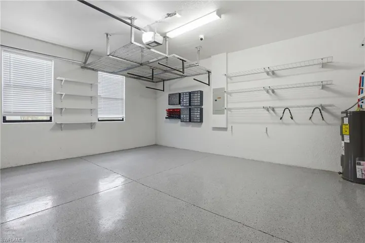Garage interior featuring a speckled epoxy floor coating, two windows with blinds, wall-mounted shelving, overhead storage racks, and a water heater