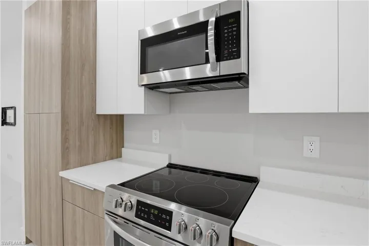Modern kitchen featuring a stainless steel range with a flat cooktop, an over-the-range microwave, white upper cabinetry, light wood-finish lower cabinetry, and white countertops