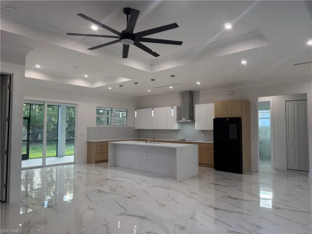 Kitchen featuring wall chimney range hood, a kitchen island with sink, backsplash , appliances, white cabinets, and a raised ceiling