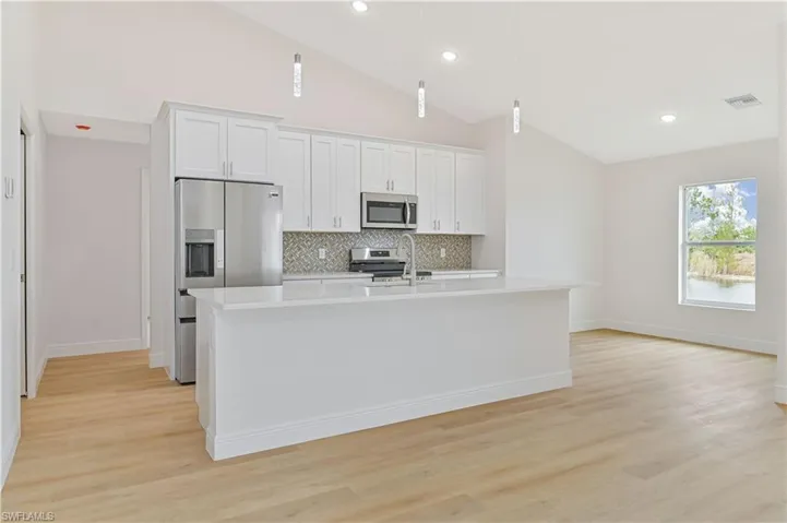 Kitchen featuring white cabinets, stainless steel appliances, a center island with sink, light wood finished floors, and lofted ceiling