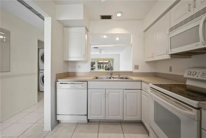 Kitchen with light tile patterned floors, white cabinetry, sink, stacked washer / dryer, and white appliances