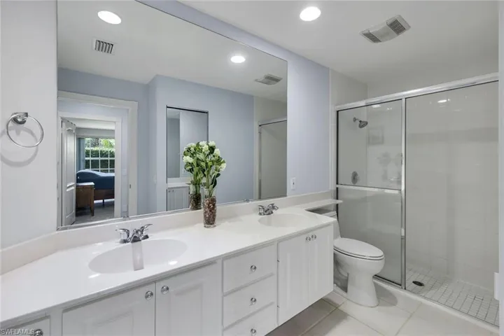 Dual sink vanity with white cabinetry and chrome fixtures, frameless mirror, recessed lighting, and a glass-enclosed shower with a sliding door