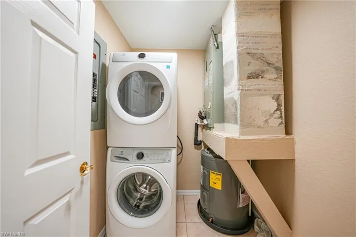 Laundry area featuring a textured wall, tile patterned flooring, water heater, stacked washer and clothes dryer, and electric panel