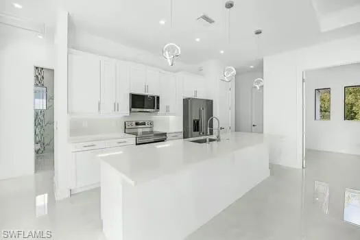 Kitchen with white cabinets, stainless steel appliances, an island with sink, hanging light fixtures, and concrete flooring