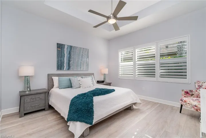 Primary bedroom with plantation shutters, LVT flooring, tray ceiling, and ceiling fan.