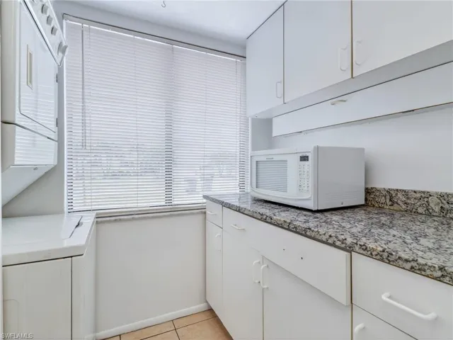 Compact utility area featuring a stacked washer and dryer unit, white cabinetry, a large window with horizontal blinds, and a speckled countertop