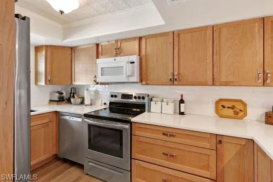 Kitchen with appliances with stainless steel finishes, light countertops, a raised ceiling, light brown cabinets, and ornamental molding