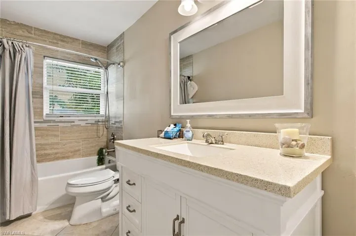 White vanity with a neutral-toned solid surface countertop and undermount sink, complemented by a large framed mirror, wall-mounted lighting, and a bathtub surround featuring horizontally-set tile