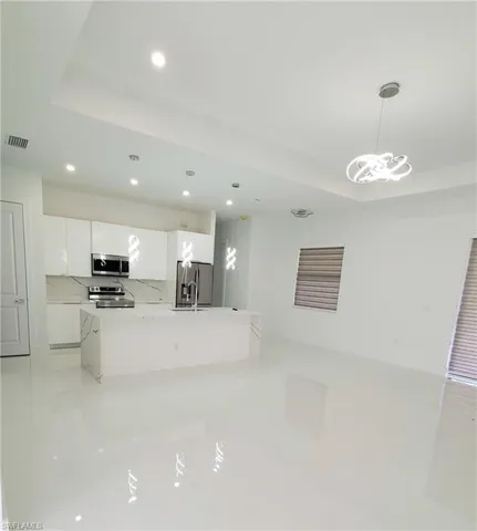 Kitchen with backsplash, a kitchen island with sink, white cabinets, appliances with stainless steel finishes, and decorative light fixtures