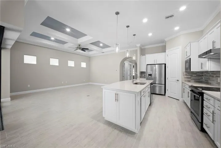 Kitchen with arched walkways, white cabinetry, stainless steel appliances, coffered ceiling, and a kitchen island with sink
