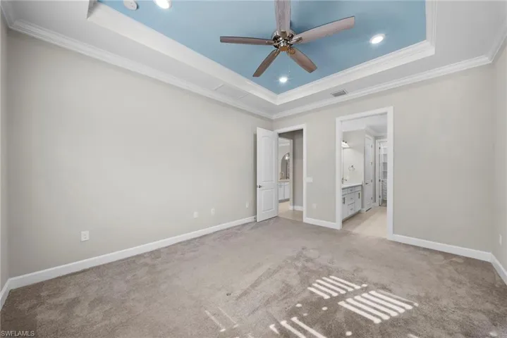 Unfurnished bedroom featuring light carpet, a raised ceiling, crown molding, ceiling fan, and connected bathroom
