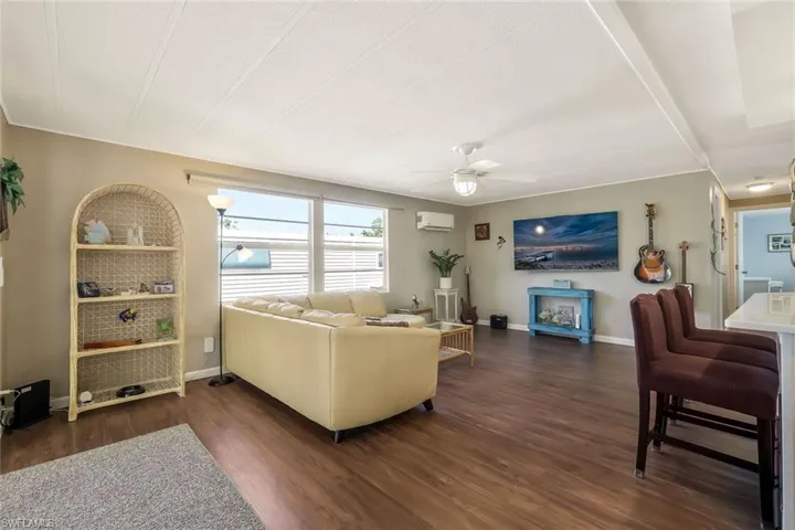 Living area with dark wood-style floors, a wall unit AC, ceiling fan, and baseboards