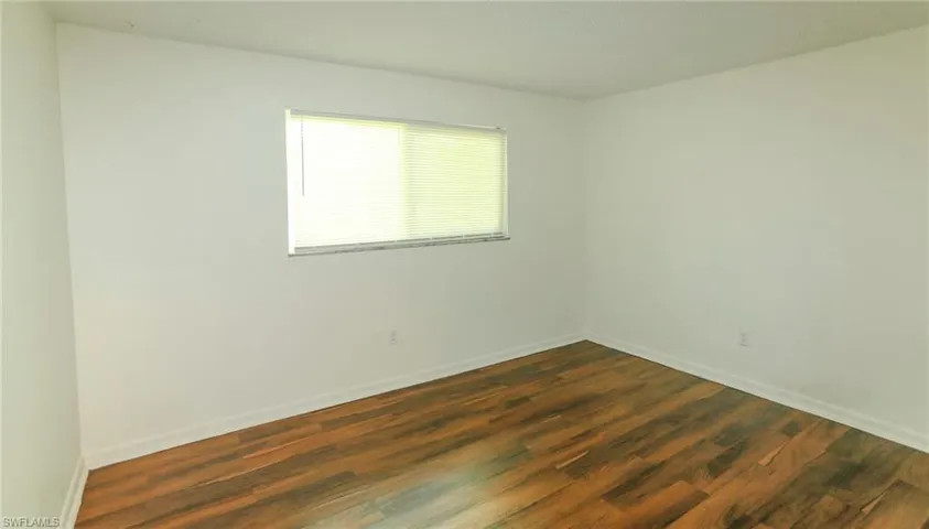 Wood-finish flooring with white baseboards, a rectangular window with blinds, and white painted walls