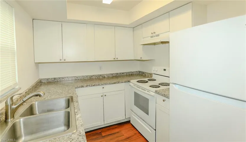 White cabinetry, speckled countertops, wood-finish flooring, double basin sink, and white appliances