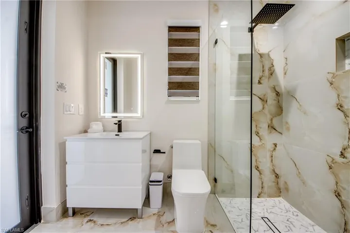 Modern bathroom featuring a glass-enclosed shower with a matte black rain showerhead, a wall-mounted LED-backlit mirror, a white vanity with a black faucet, a contemporary toilet, and patterned tile flooring