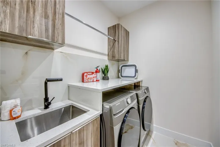 Dedicated laundry area featuring a stainless steel sink with a matte black faucet, integrated countertop, wood-finish cabinetry, and contemporary floor tiling