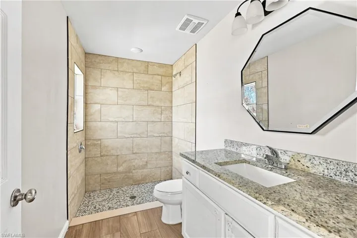 Tiled walk-in shower with pebble floor, wood-finish flooring, white vanity with granite-style countertop, undermount sink, and an octagonal mirror