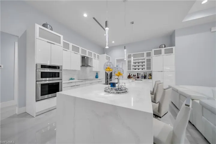 Contemporary kitchen featuring a waterfall island with a light-toned countertop, built-in double ovens, white high-gloss cabinetry, stainless steel appliances, and recessed lighting