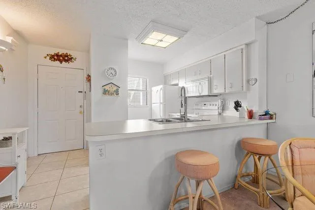Kitchen featuring kitchen peninsula, white appliances, a textured ceiling, light tile floors, and a kitchen bar