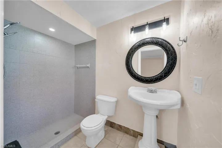 Bathroom featuring a walk-in shower with gray tile surround, pedestal sink with a decorative round mirror, and a contemporary vanity light fixture