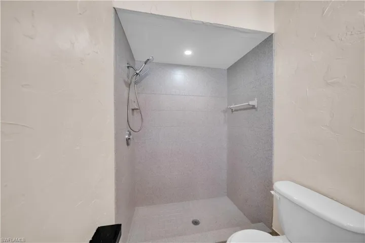 Recessed shower with light gray tile walls and flooring, featuring a handheld showerhead and built-in bench