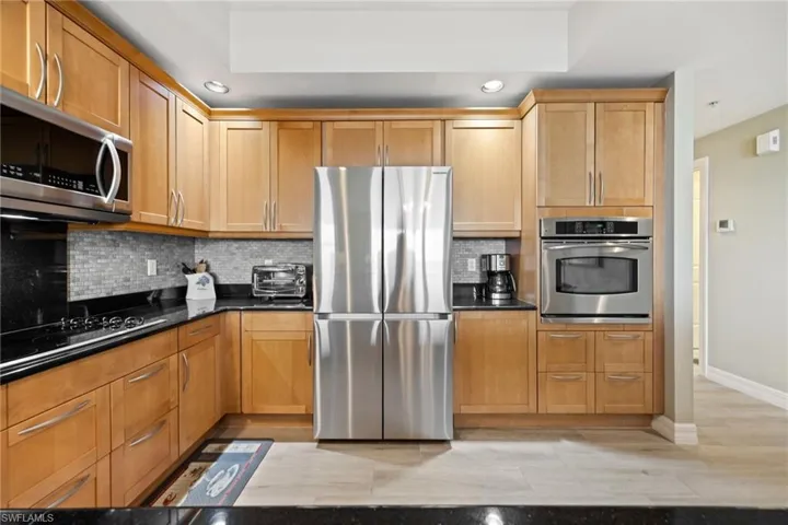 Kitchen with Newer stainless-steel Fridge, recessed lighting, wood cabinetry and dark granite counters with backsplash.