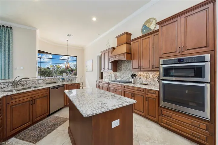 Kitchen featuring brown cabinetry, stainless steel appliances, light stone counters, ornamental molding, and recessed lighting