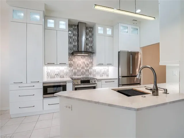 Kitchen with wall chimney range hood, white cabinetry, light stone counters, kitchen peninsula, and stainless steel appliances