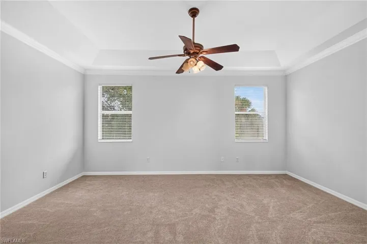 Tray ceiling with crown molding, ceiling fan with light fixture, dual windows with blinds, and neutral carpeting