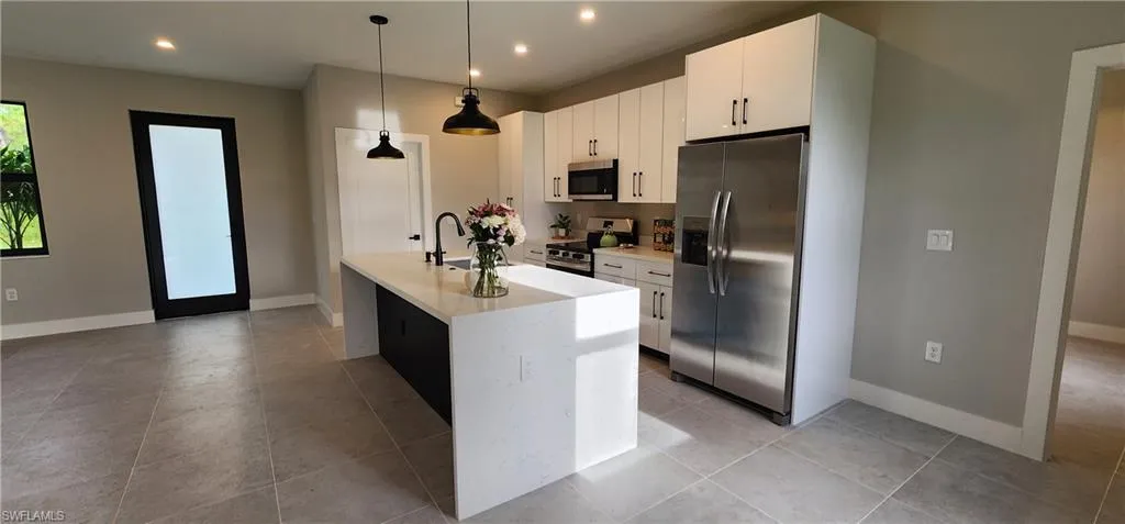 Kitchen featuring appliances with stainless steel finishes, hanging light fixtures, light stone countertops, white cabinetry, and a kitchen island with sink