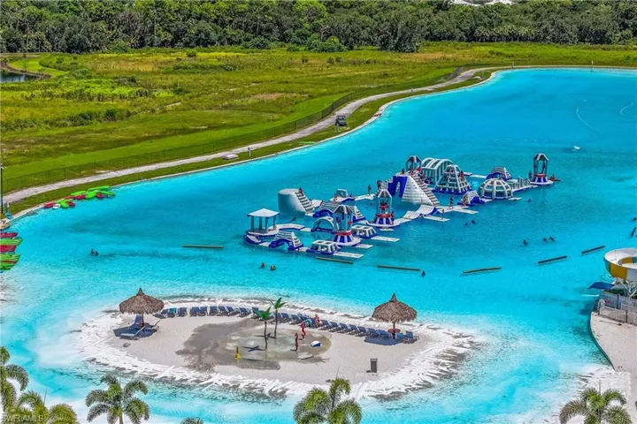 Expansive blue water lagoon with a large inflatable obstacle course, private sandy island with thatched umbrellas, and shoreline recreational equipment