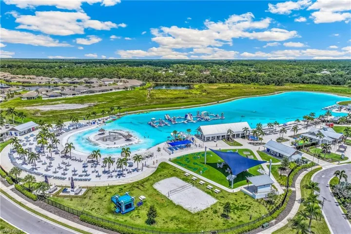 Expansive community amenity area featuring a large lagoon with clear blue water, a sandy beach area with numerous lounge chairs, multiple recreational buildings, and a dedicated volleyball court
