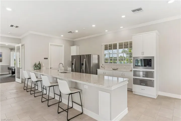Kitchen island with a light-colored countertop and integrated sink, stainless steel appliances, white cabinetry with built-in microwave, light-toned tile flooring, and recessed lighting