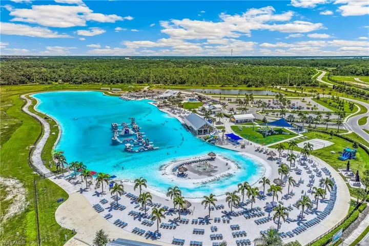 Expansive lagoon with aqua park features, surrounding sandy beach area, multiple lounge chairs, mature palm trees, and adjacent recreation building