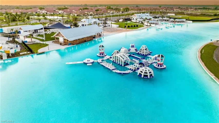 Expansive clear water lagoon featuring an inflatable obstacle course, adjacent sandy beach area, and multiple waterside structures with covered outdoor spaces