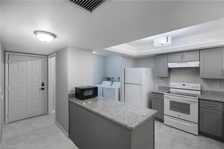 Kitchen with gray cabinetry, white appliances, washer and dryer, light stone counters, and a peninsula