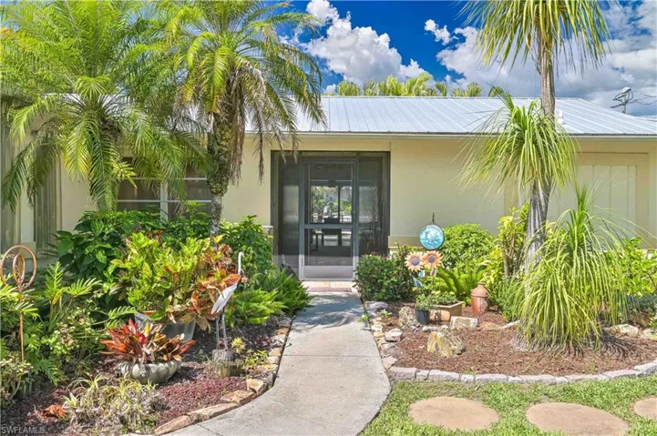 The property features a well-maintained front yard with lush landscaping, palm trees, and a paved walkway leading to the screened entry