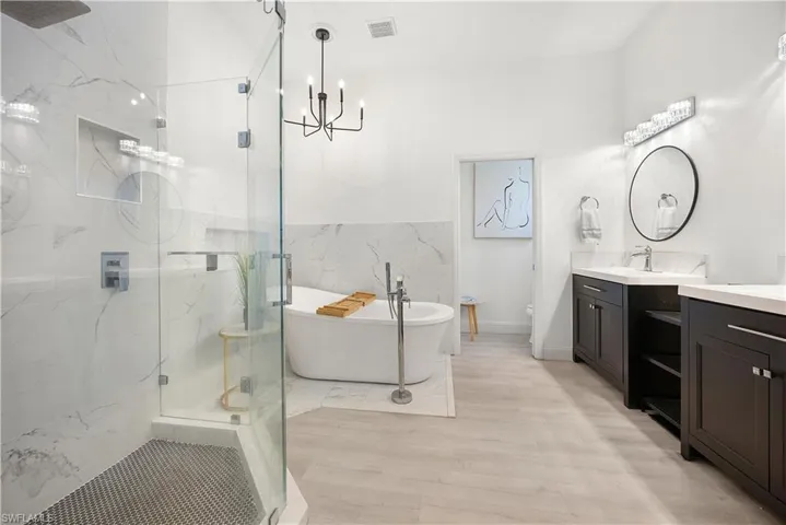 Contemporary bathroom featuring a freestanding soaking tub, clear glass shower enclosure with hex tile flooring, dual dark wood vanities with white countertops, and wood-finish flooring