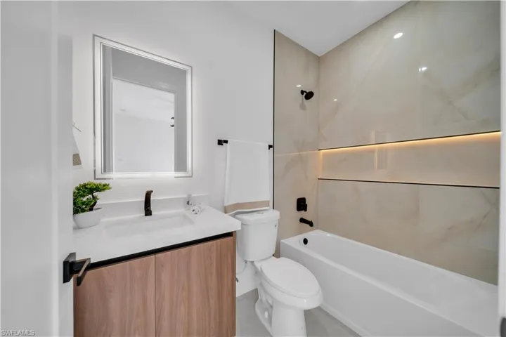 Contemporary bathroom featuring a wood-finish vanity, integrated sink with matte black fixtures, an illuminated mirror, a white bathtub, and large format wall tile with accent lighting