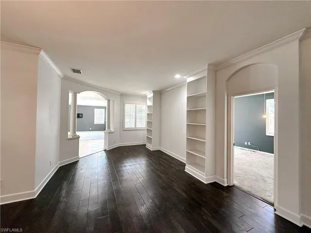 Unfurnished living room featuring arched walkways, decorative columns, dark wood-style floors, ornamental molding, and built in shelves