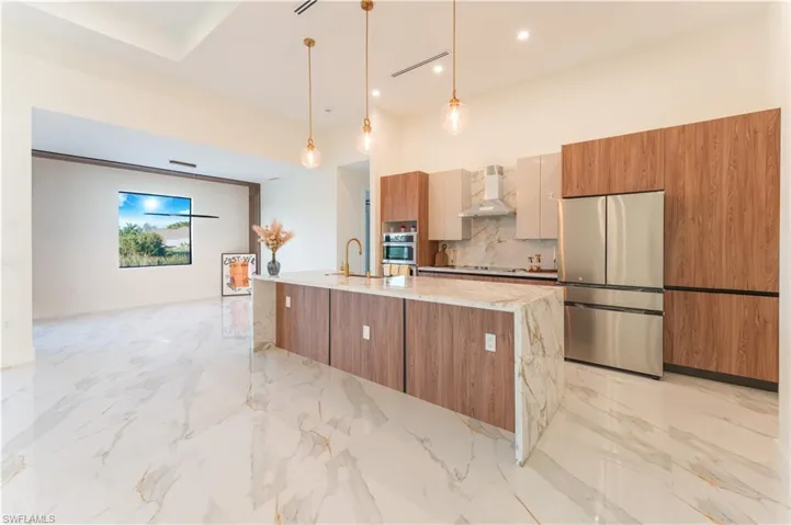 Kitchen featuring wall chimney exhaust hood, hanging light fixtures, light stone counters, a large island with sink, and appliances with stainless steel finishes
