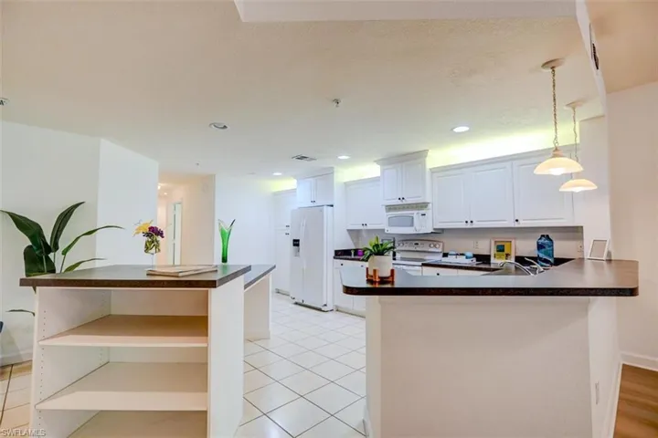 Kitchen featuring light tile patterned flooring, hanging light fixtures, kitchen peninsula, white appliances, and white cabinetry