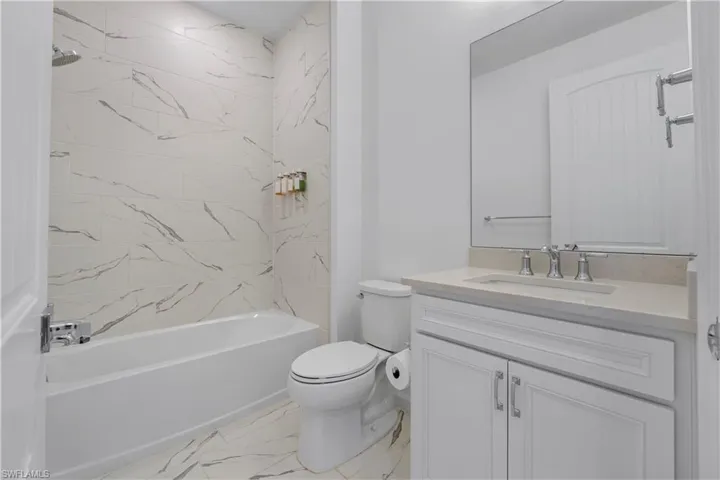White bathtub with chrome fixtures, large format white and gray veined tile surround, vanity with white shaker-style cabinetry, undermount sink, and light-toned countertop