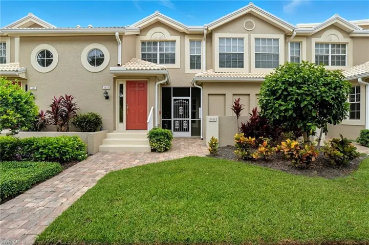 Stucco exterior with a tiled roof, featuring a vibrant red entry door, a brick paver walkway, and lush landscaping