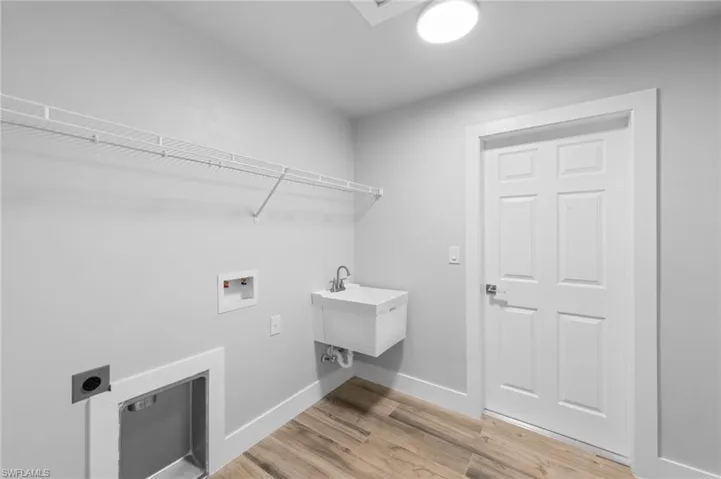 Laundry area featuring wood-finish flooring, a utility sink with a chrome faucet, a six-panel door, light gray walls, and an overhead wire shelf