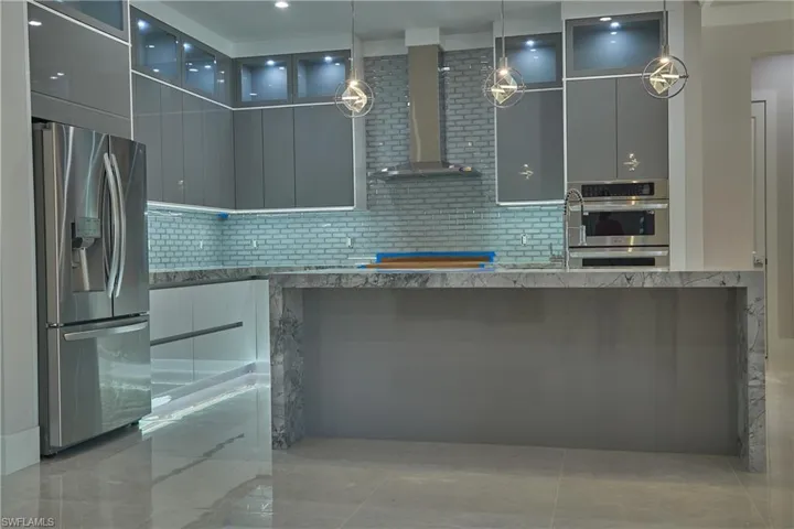 Kitchen with light tile patterned floors, wall chimney range hood, gray cabinets, appliances with stainless steel finishes, and decorative backsplash