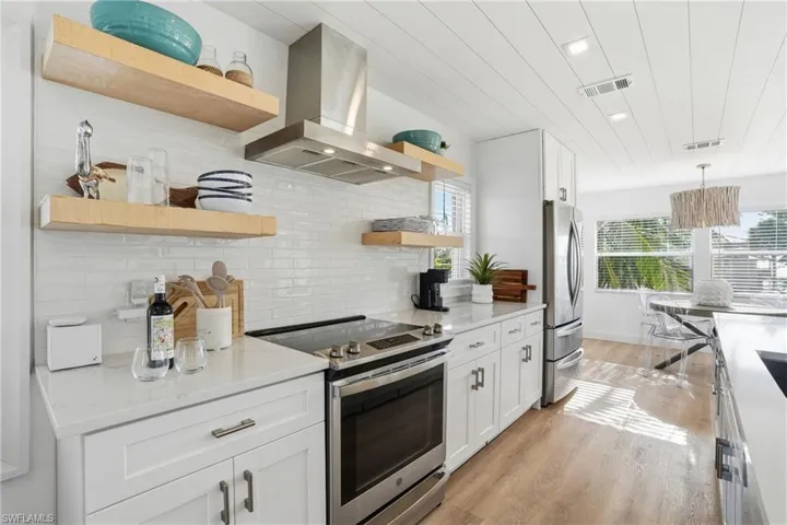 Kitchen with open shelves, appliances with stainless steel finishes, white cabinetry, wall chimney range hood, and light stone counters Virtually Edited
