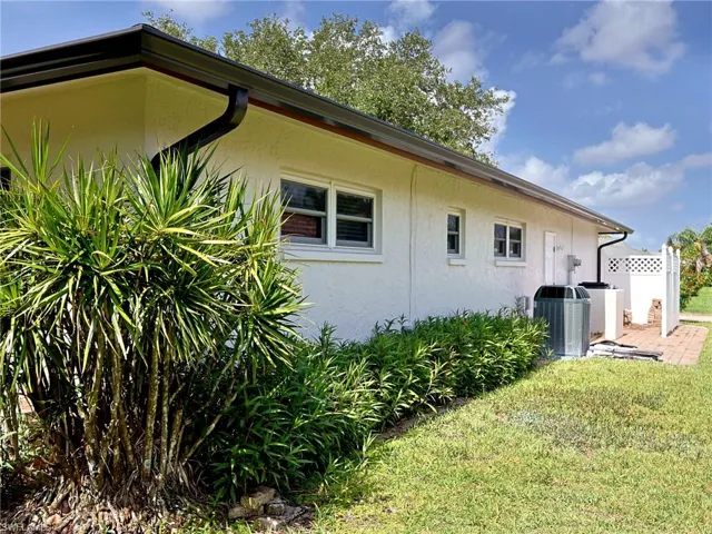White stucco exterior with black gutters and downspouts, featuring multiple windows, a robust air conditioning unit, and a section of white lattice fencing
