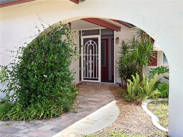 Arched stucco entryway featuring a paver walkway, mature shrubbery, decorative security screen door, and a red front door